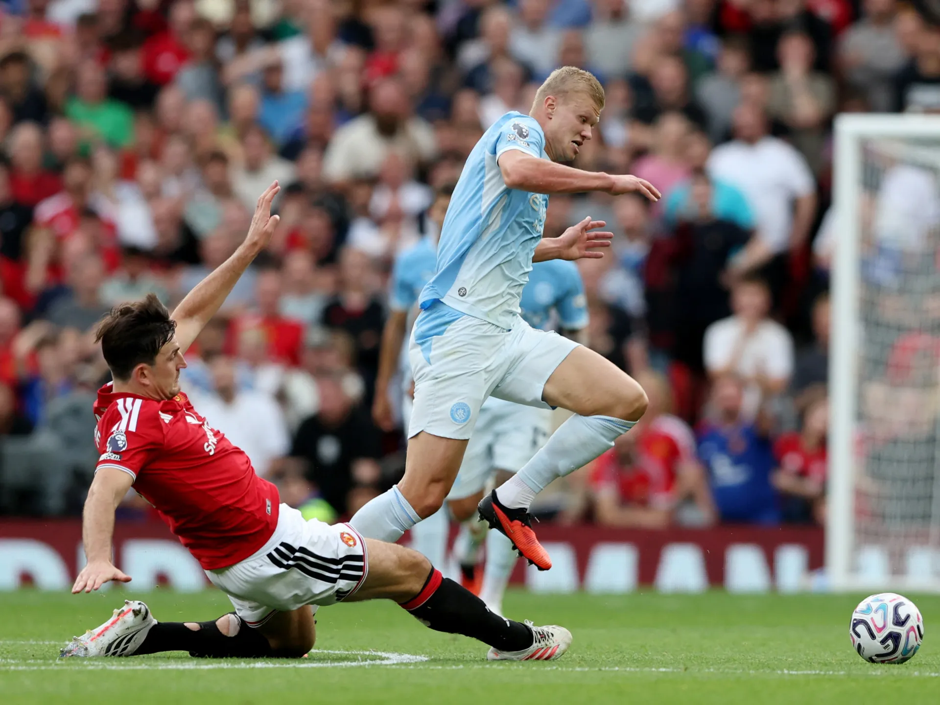 Footballer in red slides as a light-blue-shirted player runs toward the ball near goal