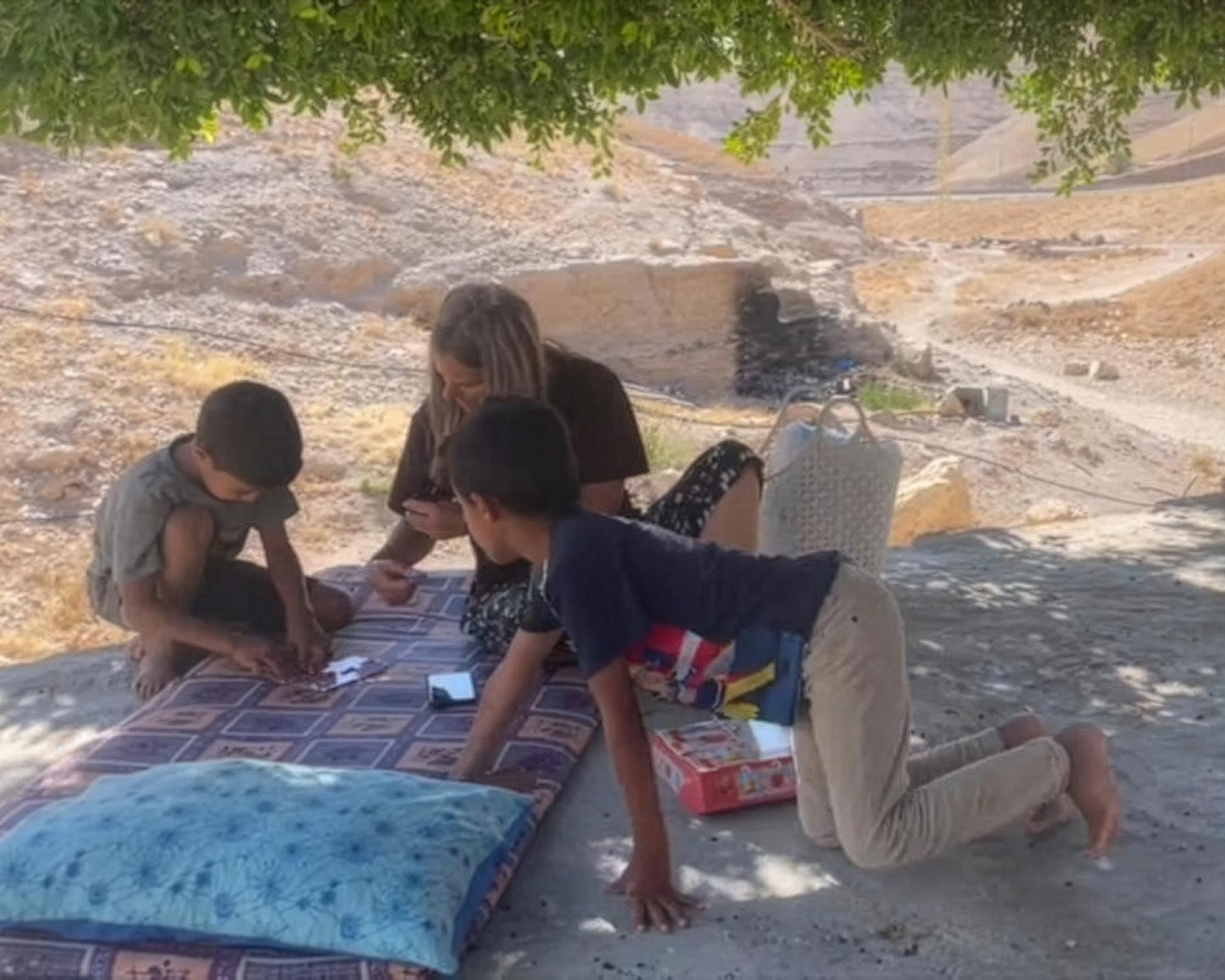 Four people sit outdoors on a patterned mat beneath a tree near a barren construction site.