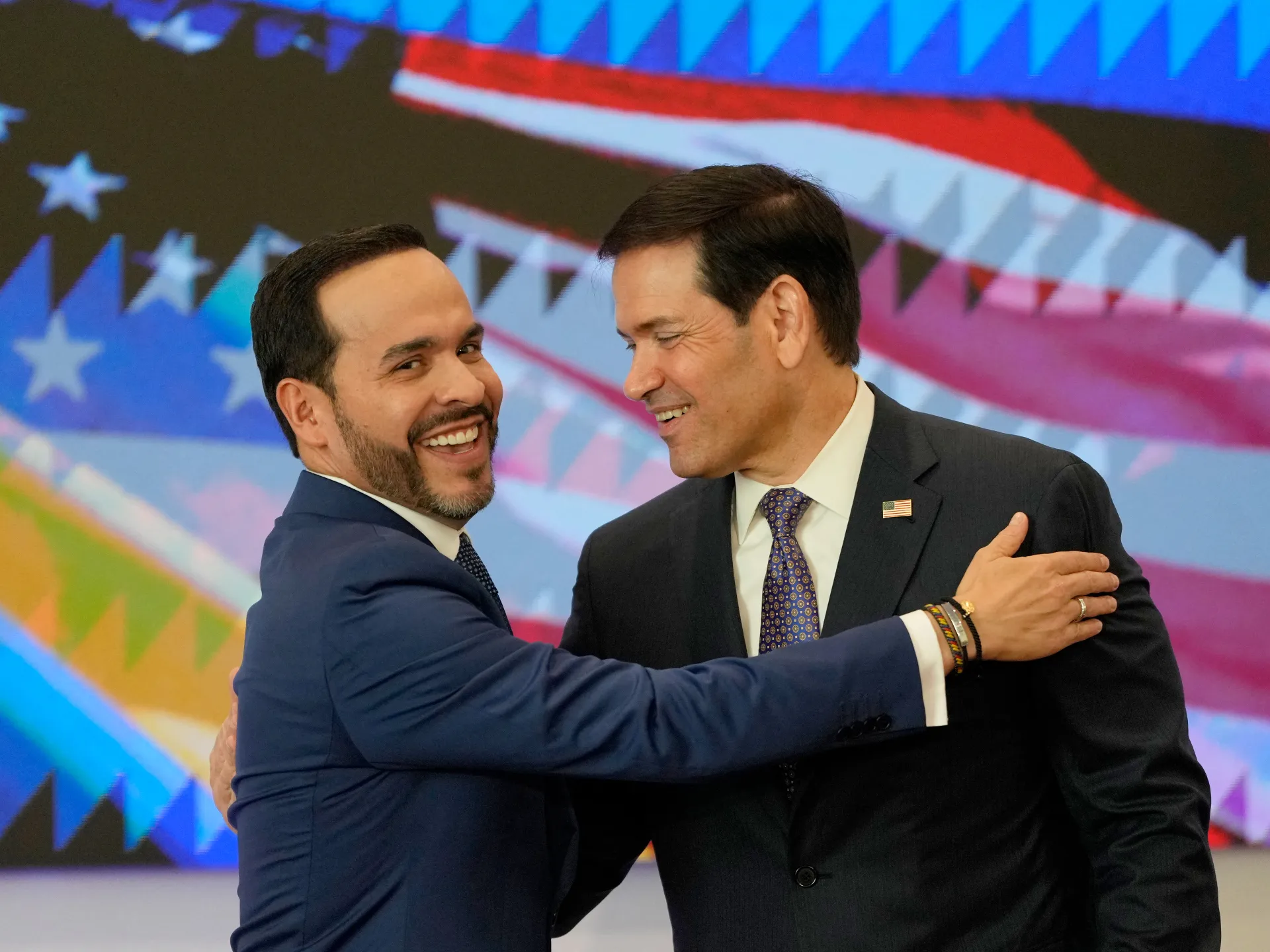 Two suited men smile and embrace before a large American flag display.
