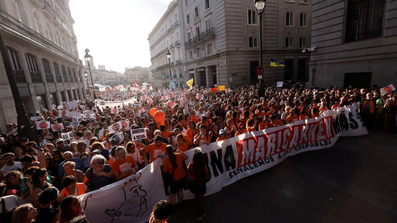 A dense street demonstration with orange-clad marchers carrying signs and a large banner between city buildings.