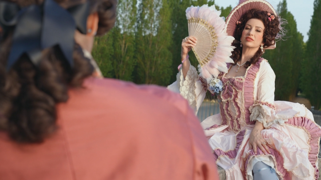 Woman in ornate pink 18th-century dress holds a feathered fan outdoors before another figure.