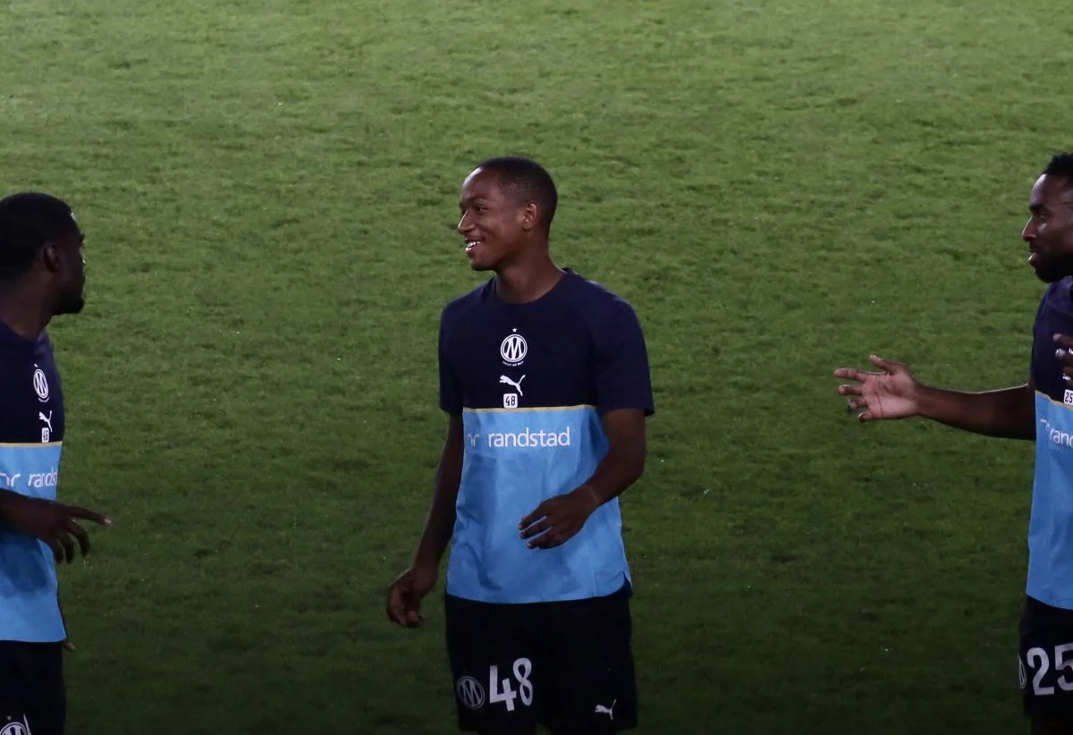 Three Marseille footballers in training tops stand and gesture on a grass pitch
