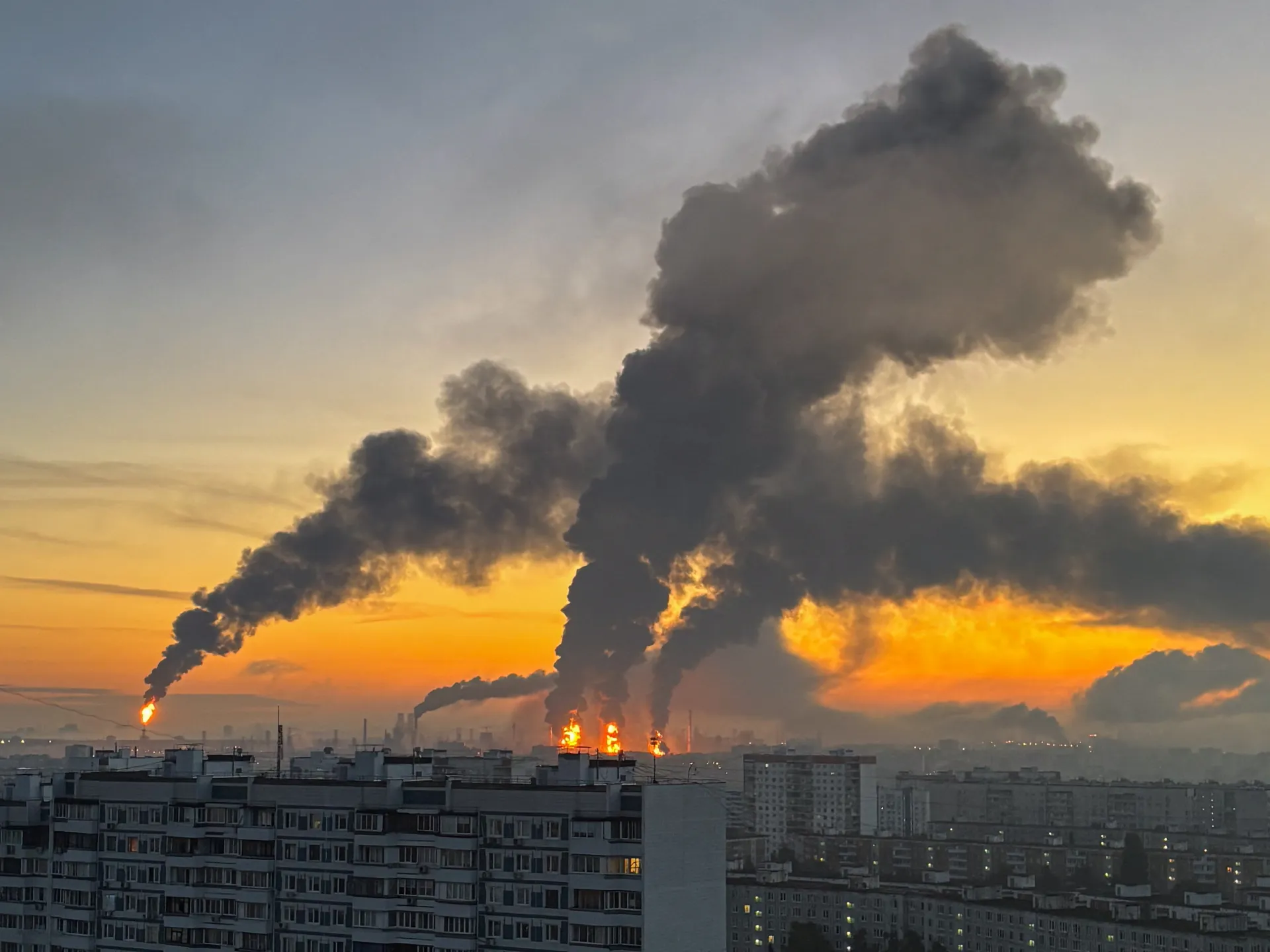 Smoke plumes and flames rise above a densely built city skyline at sunset.