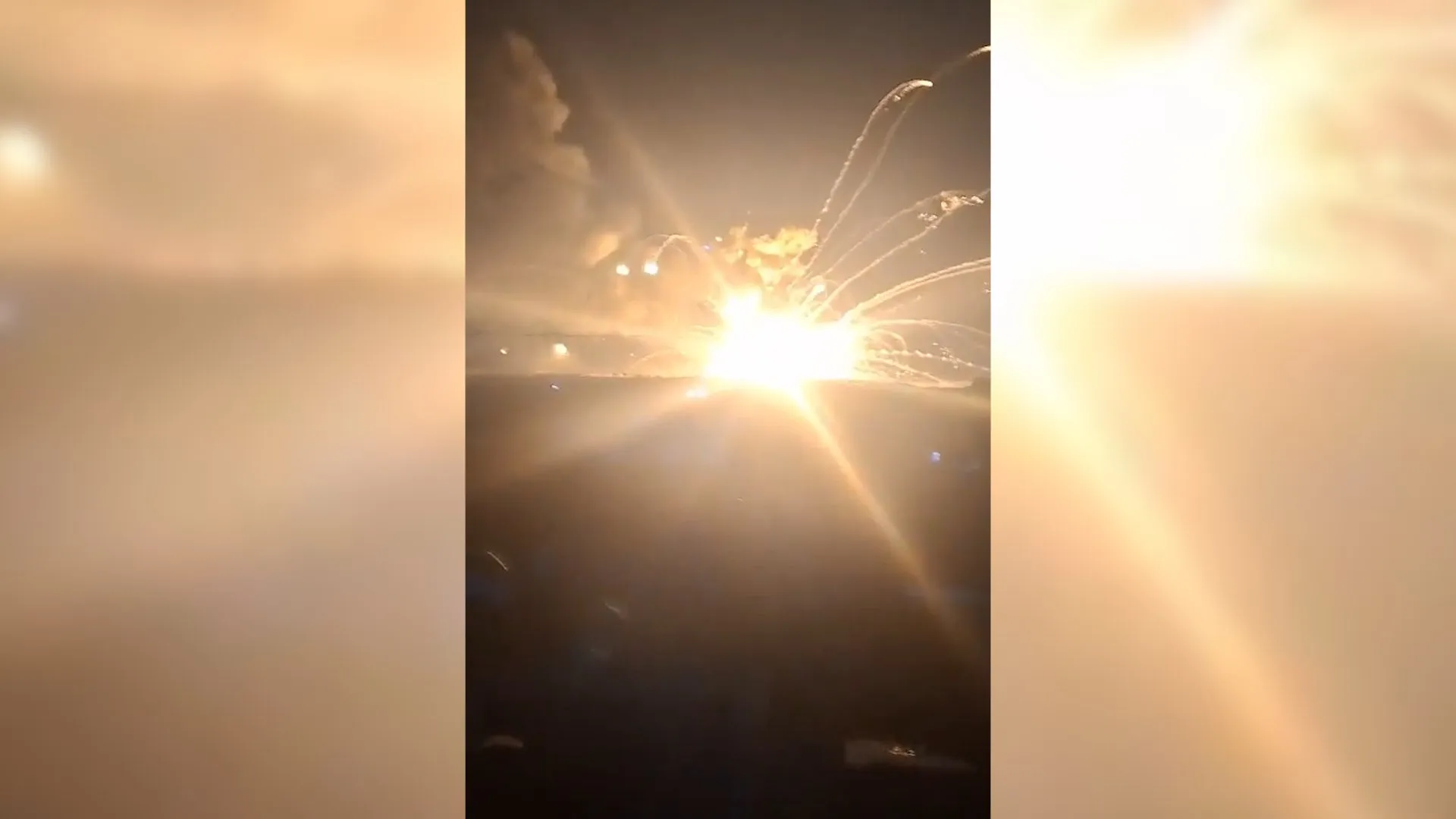 Bright explosion sends sparks and smoke into the night sky, viewed from behind a vehicle windshield.