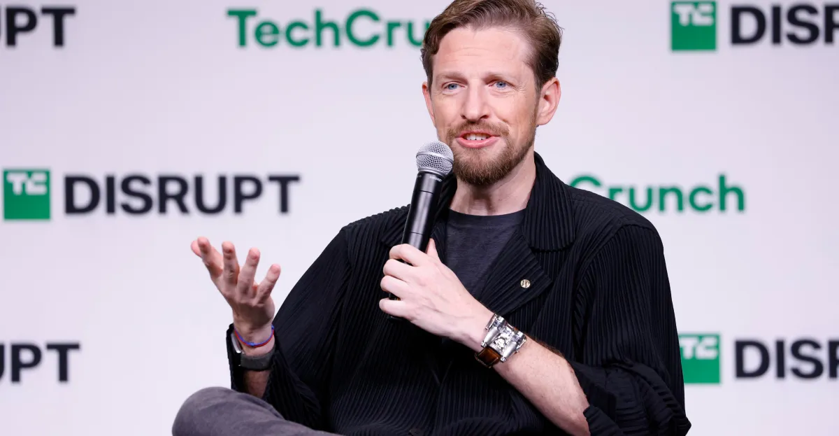 Bearded man in black clothing speaks into a microphone while gesturing onstage.
