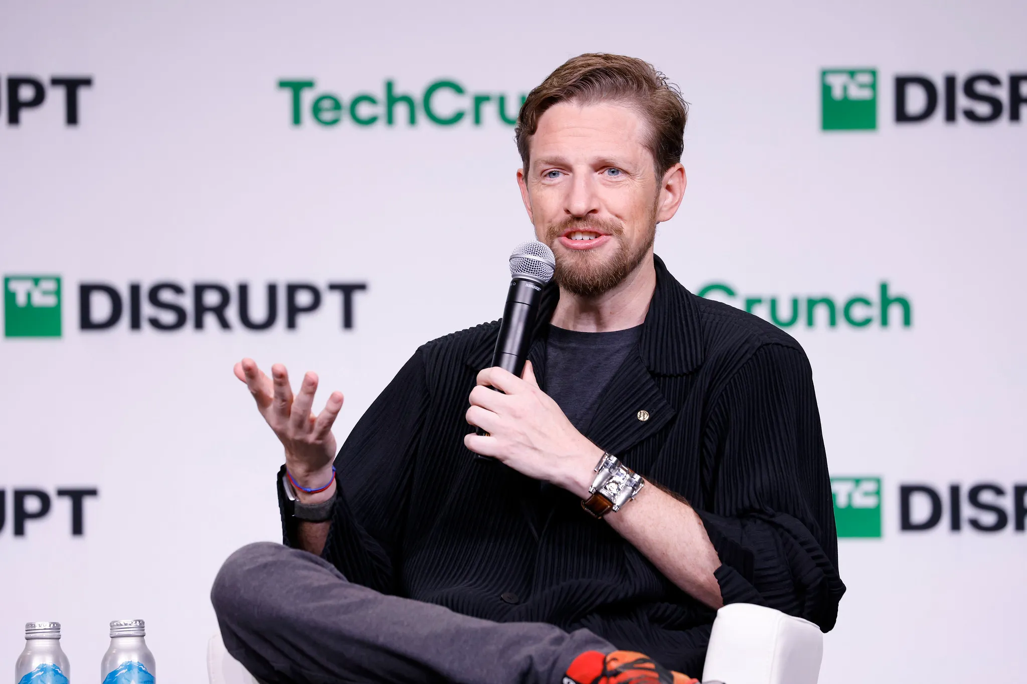 A bearded man speaks into a microphone while gesturing onstage before a branded backdrop.