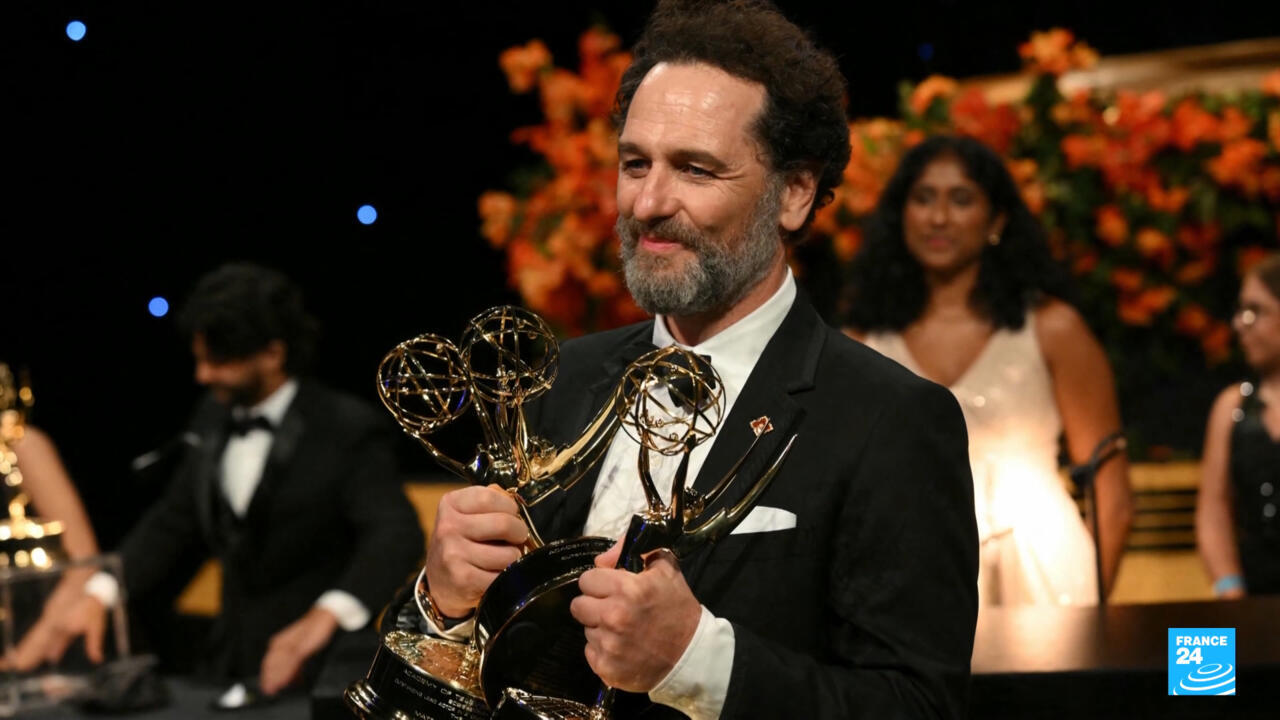 Bearded man in black tuxedo holds two Emmy statuettes during an awards ceremony.