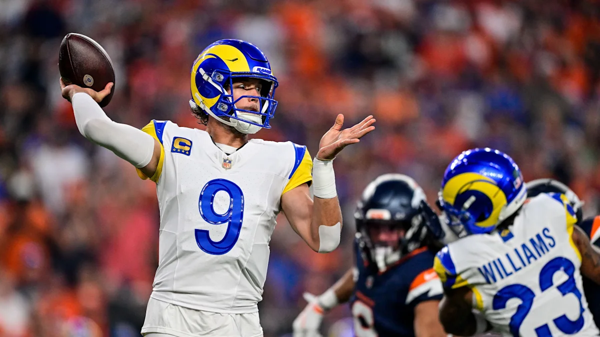 Rams quarterback in a white number 9 jersey prepares to throw as a teammate blocks a defender.