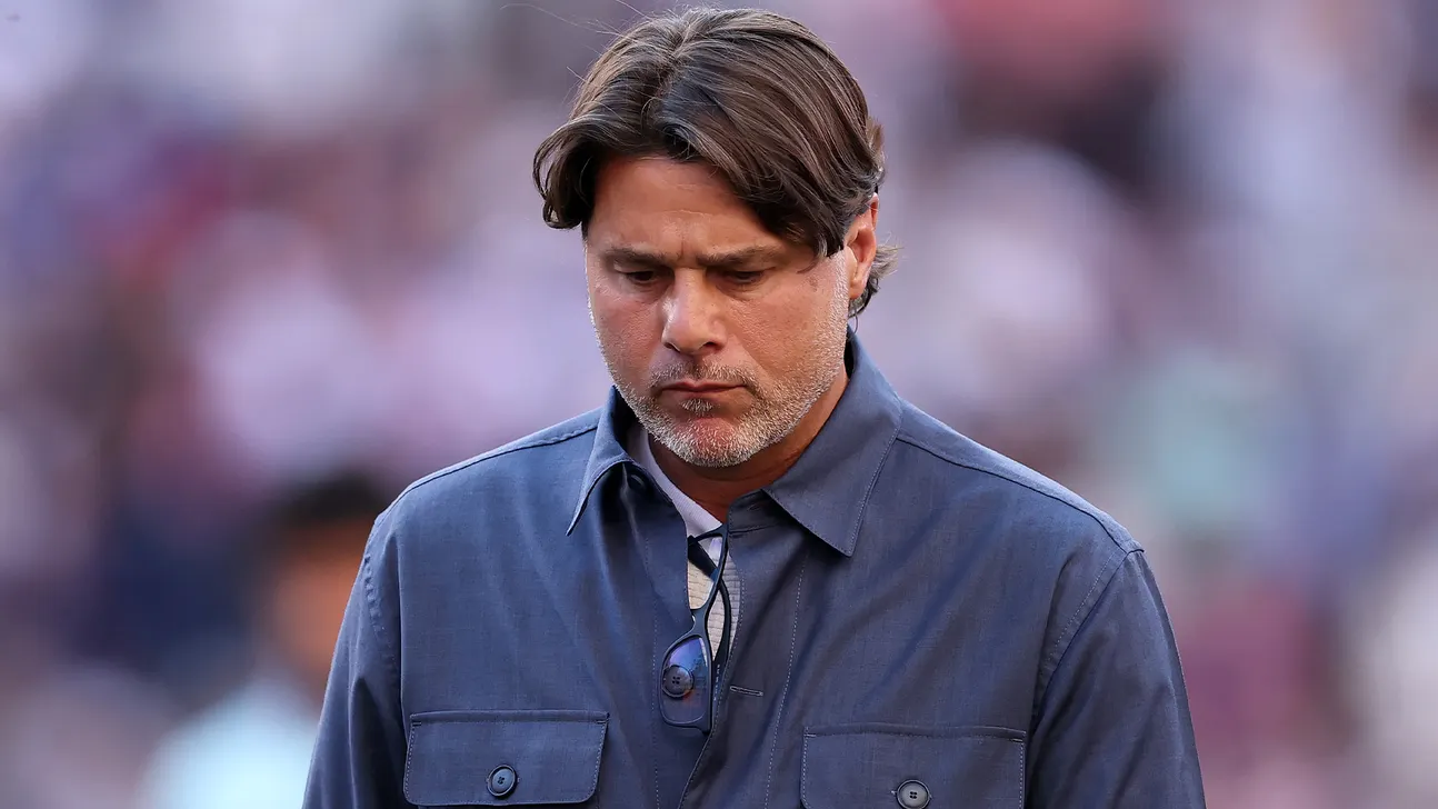 A man in a blue jacket looks down, with glasses hanging from his shirt.