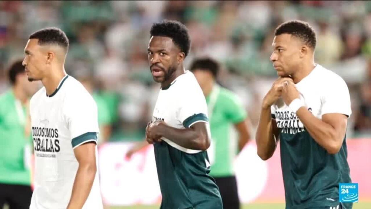 Three footballers in white and green shirts stand on a stadium pitch during a match.