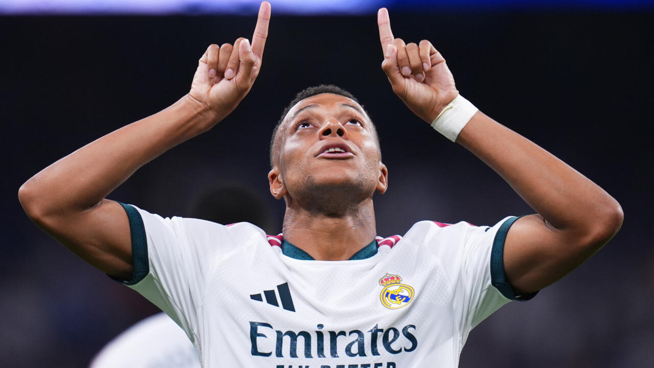 Footballer in white jersey raises both index fingers while looking upward under stadium lights.