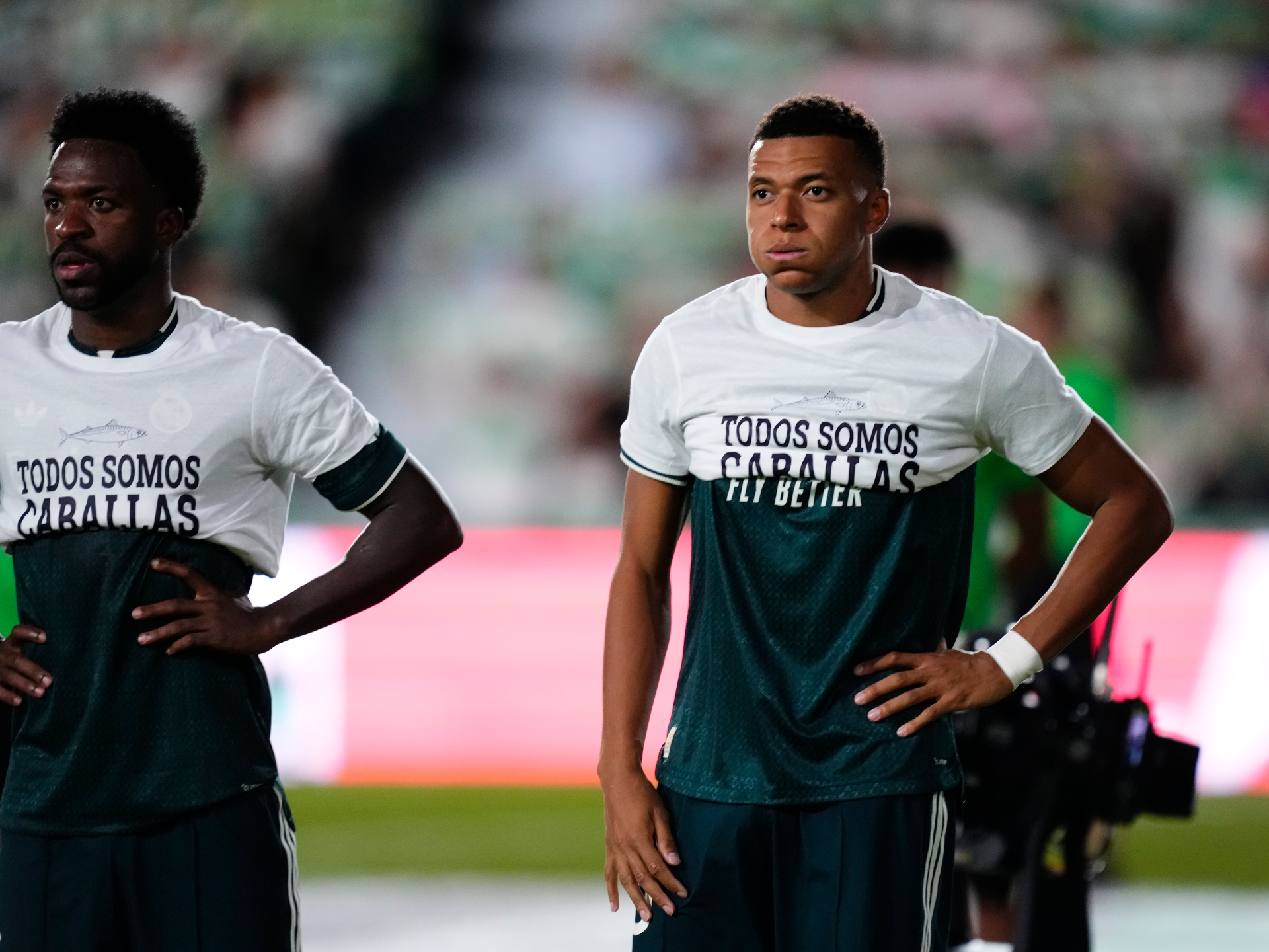Two footballers wearing white and dark shirts stand on a pitch with hands on hips.