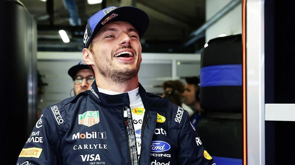 Smiling racing driver in a navy sponsor-covered suit and cap inside a garage