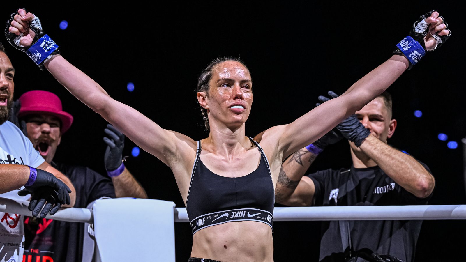 Female fighter raises both arms in victory inside a ring surrounded by cornermen