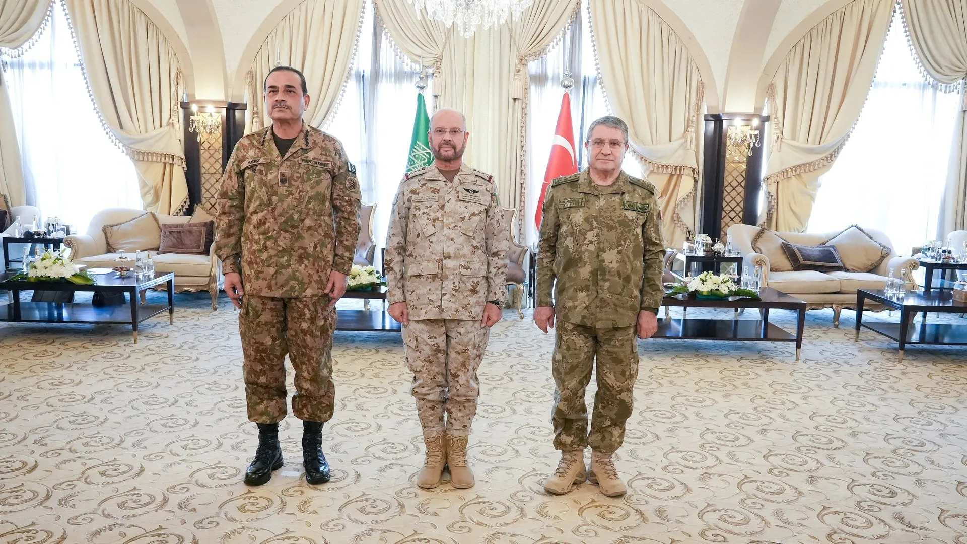 Three uniformed military officers stand indoors before Saudi and Turkish flags in a formal sitting room.