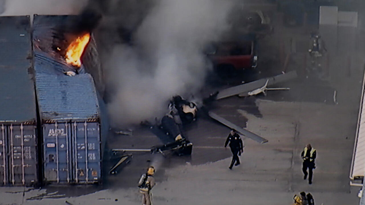 Smoke and flames rise near wreckage as emergency personnel respond beside shipping containers.