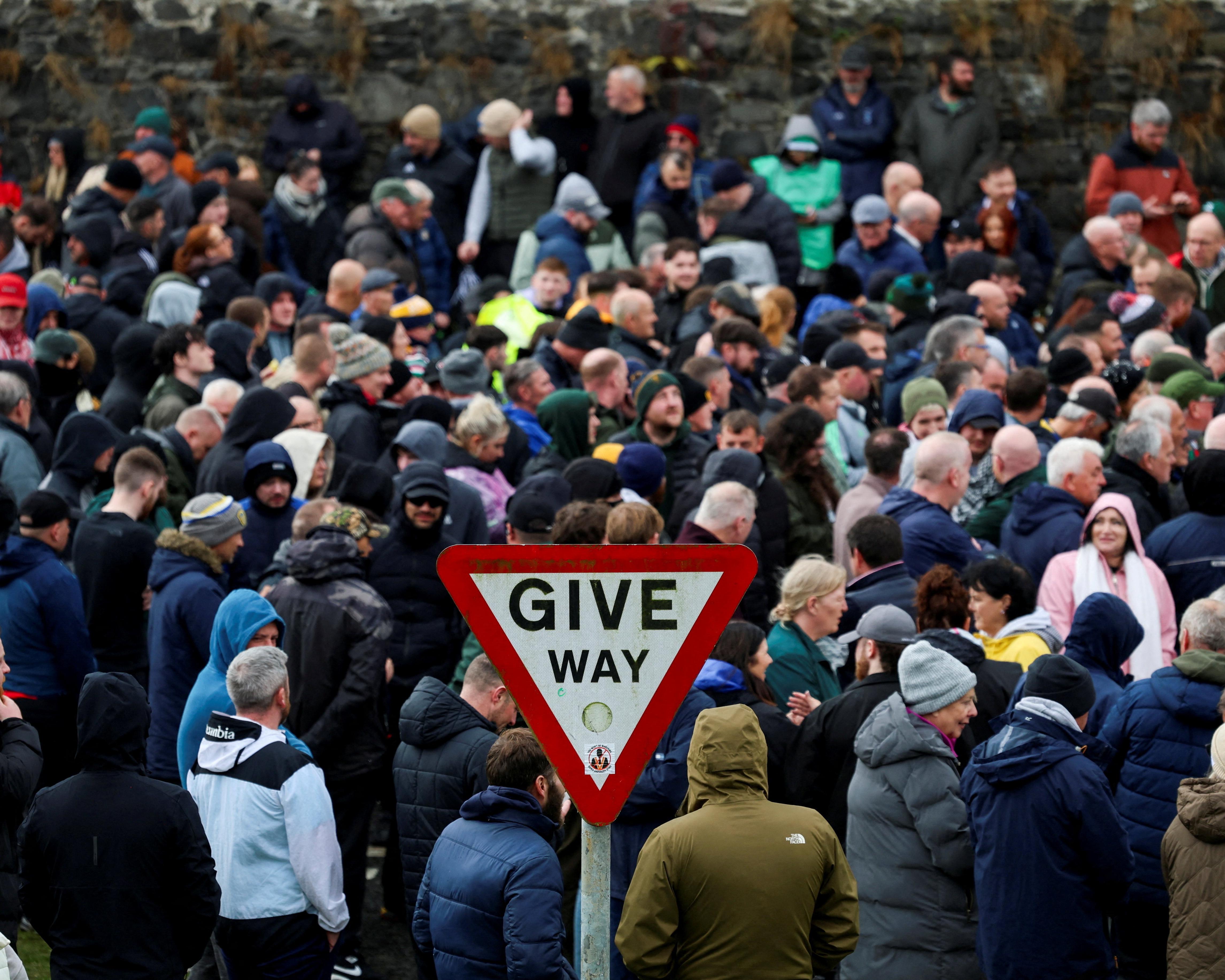 A dense crowd gathers behind a red and white give way sign, with a stone wall beyond.