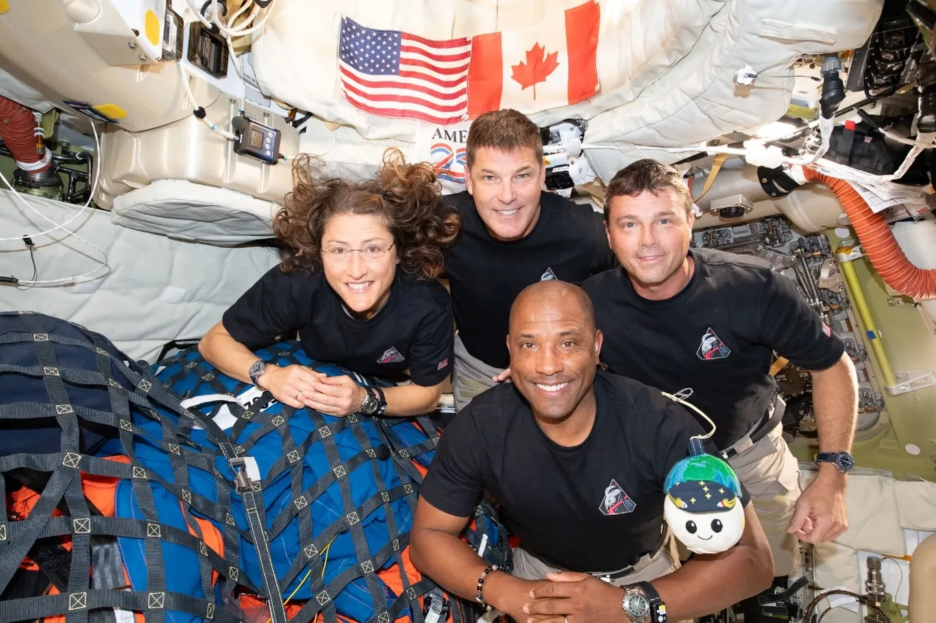 Four astronauts pose in a spacecraft cabin beneath U.S. and Canadian flags, with equipment and a small plush toy.