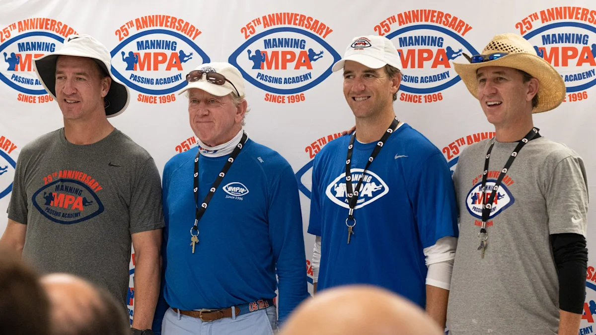 Four men wearing Manning Passing Academy shirts and hats pose before a branded backdrop.