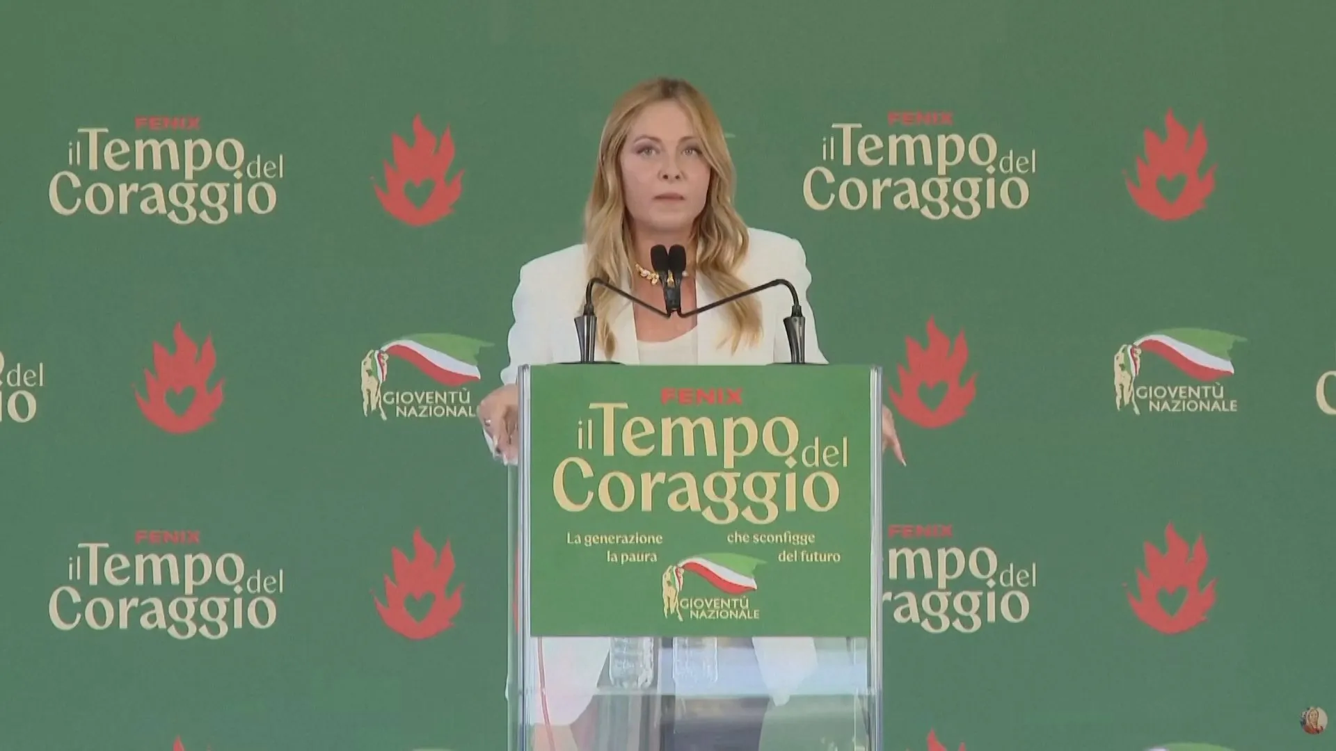 Blonde woman speaking at a podium against a green backdrop with Italian political event logos