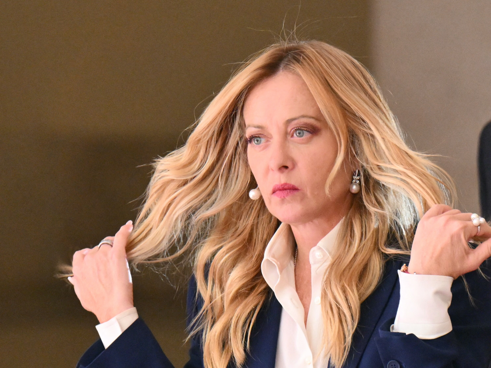 Blonde woman in a navy blazer and white shirt adjusts her hair indoors.