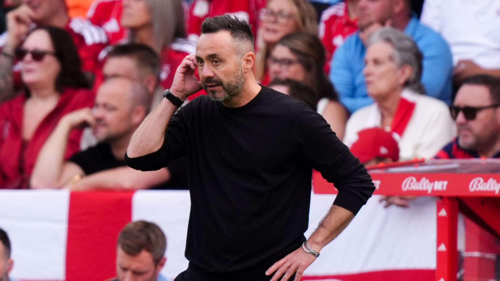 Football coach in black shirt gestures beside the pitch with supporters blurred behind him