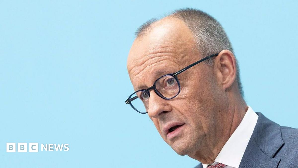 Bespectacled older man in a suit speaking against a light blue background