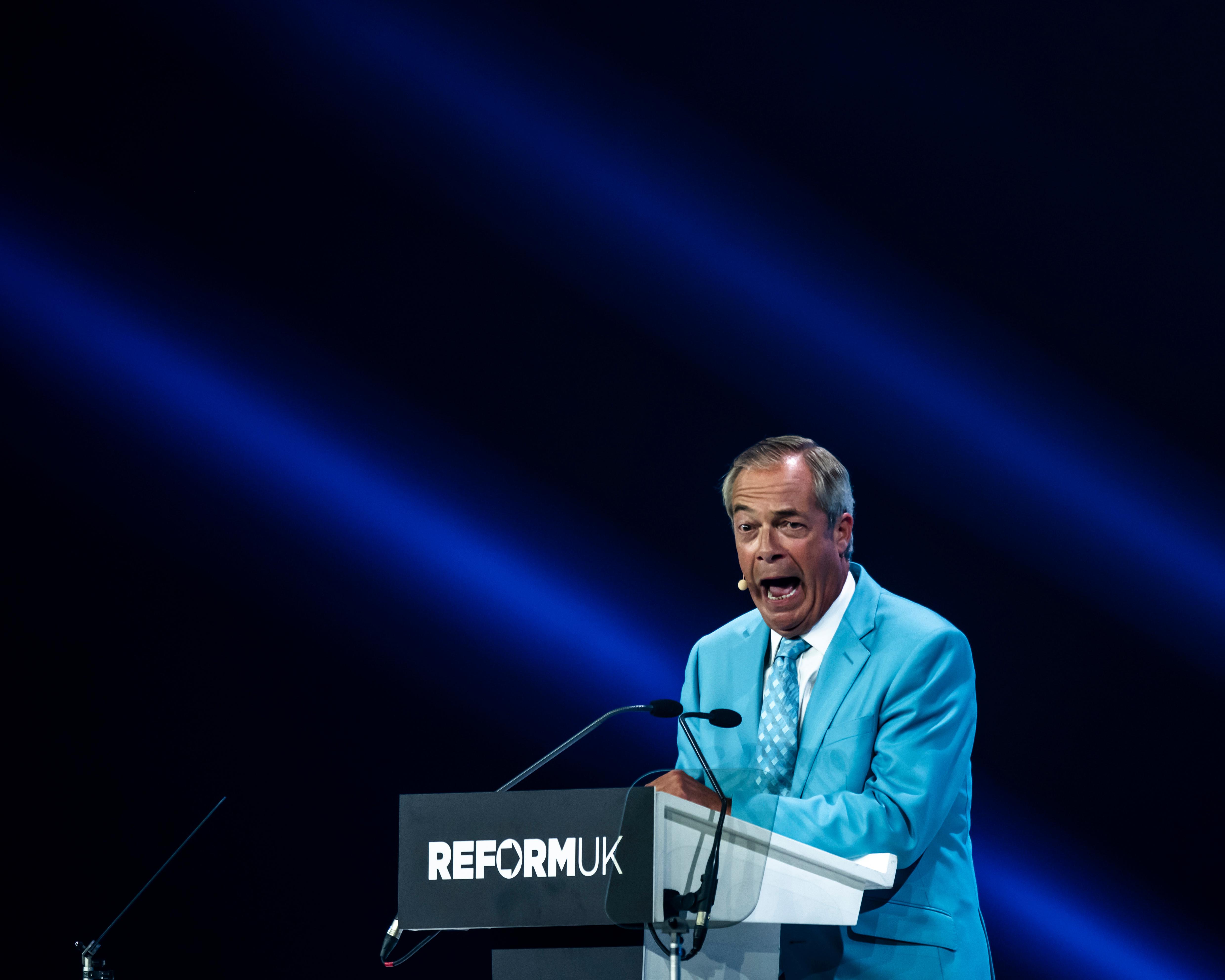 Man in turquoise suit speaking at a Reform UK lectern beneath blue stage lighting