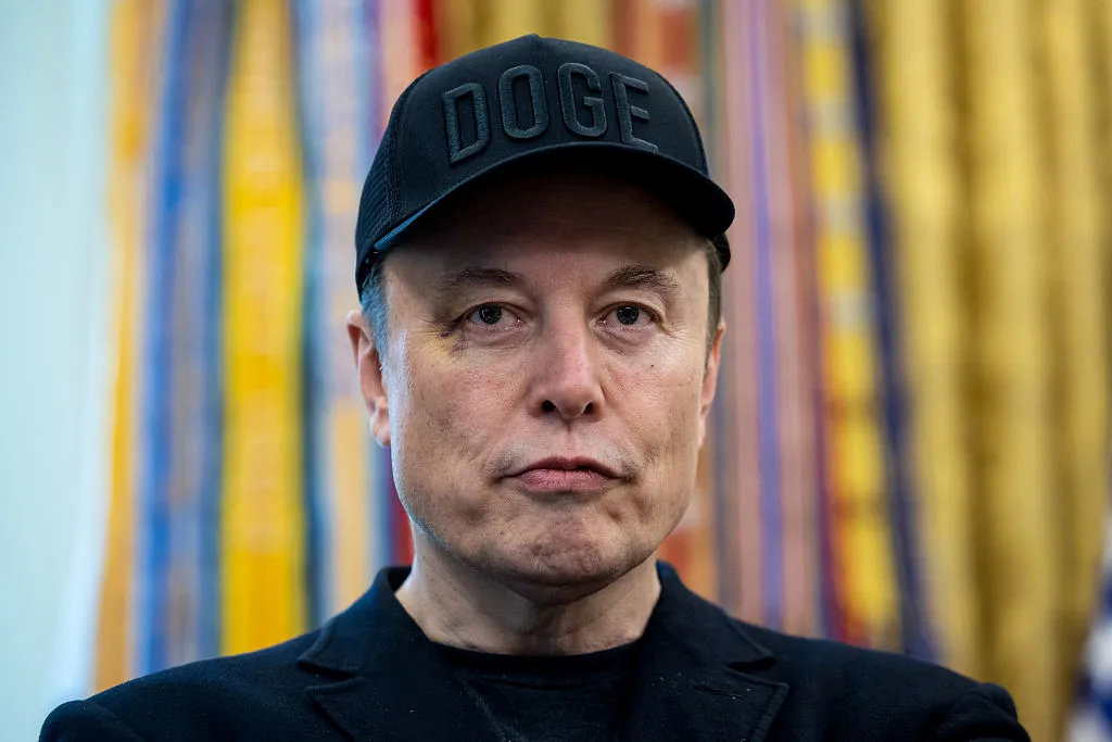 Man wearing a black DOGE cap and dark jacket, photographed against a colorful striped background.