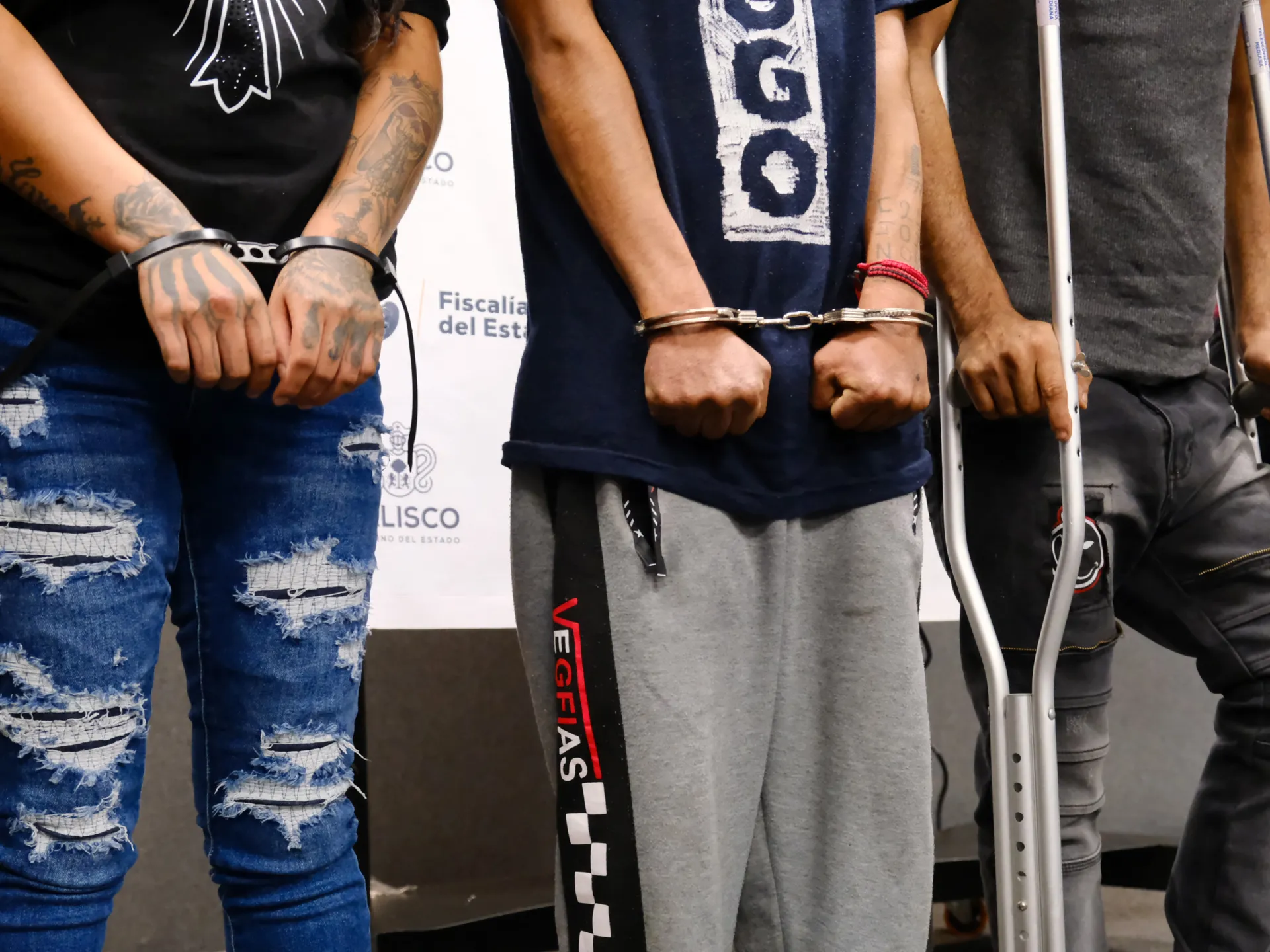 Three people stand indoors, two handcuffed, while another uses crutches beside them.