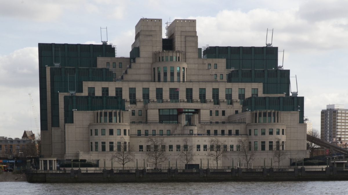 A large, tiered concrete and dark glass office building stands beside a river beneath a cloudy sky.