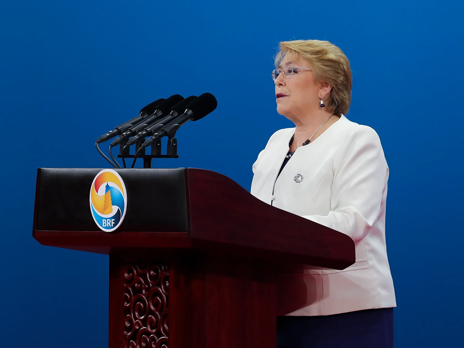 Woman in white jacket speaking at a podium with multiple microphones against a blue backdrop