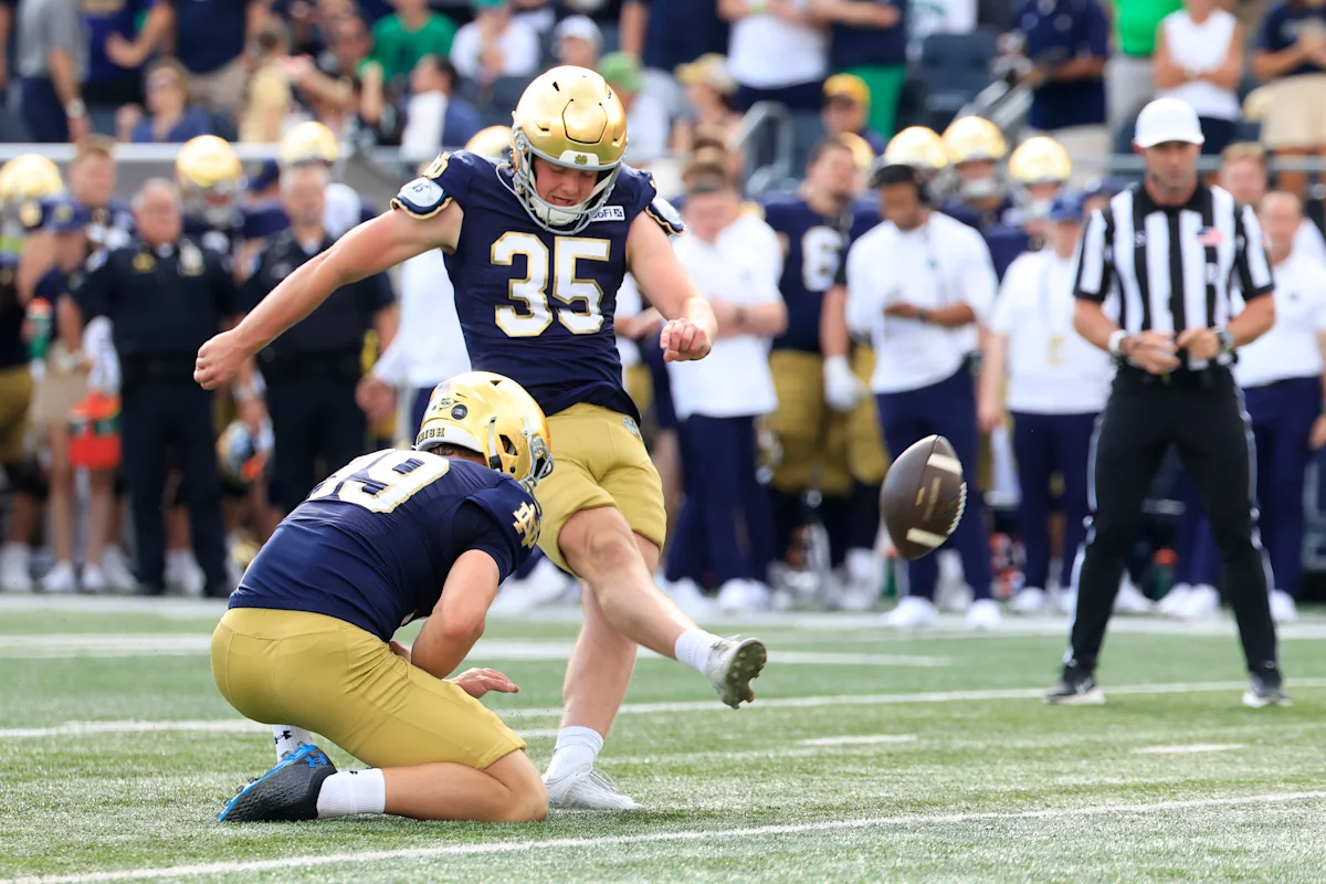 Notre Dame football player kicks a football during a game as teammate holds it