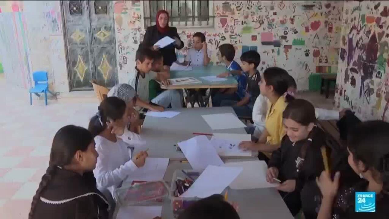 Children draw and write at tables in a brightly decorated classroom as an adult holds papers nearby.