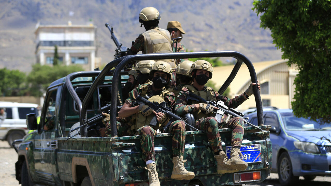 Armed soldiers in camouflage sit aboard a military pickup truck near buildings and mountains.