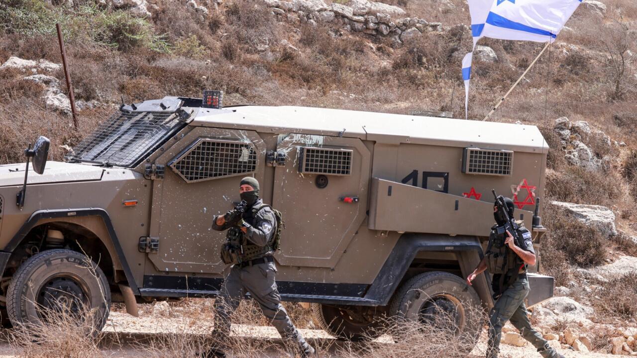Two armed personnel walk beside an armored vehicle on a rocky, dry hillside with Israeli flags visible.