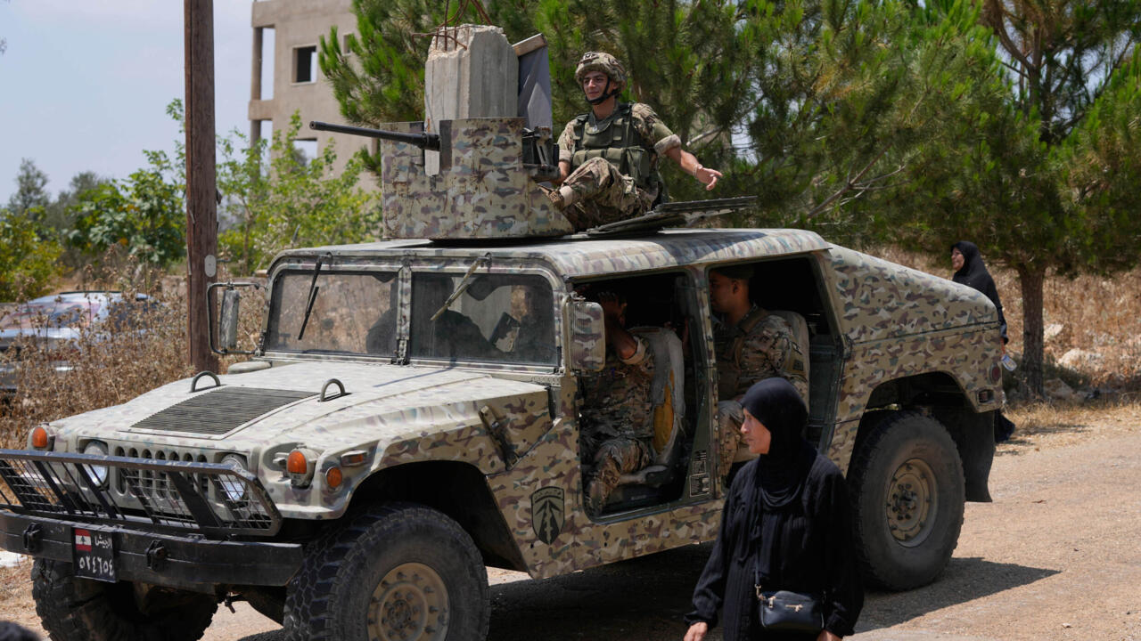 Camouflaged military vehicle with soldiers aboard and a woman standing beside it outdoors