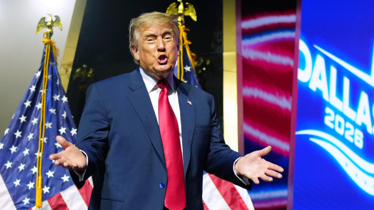 Donald Trump gestures with both hands while speaking beside U.S. flags and a campaign display.
