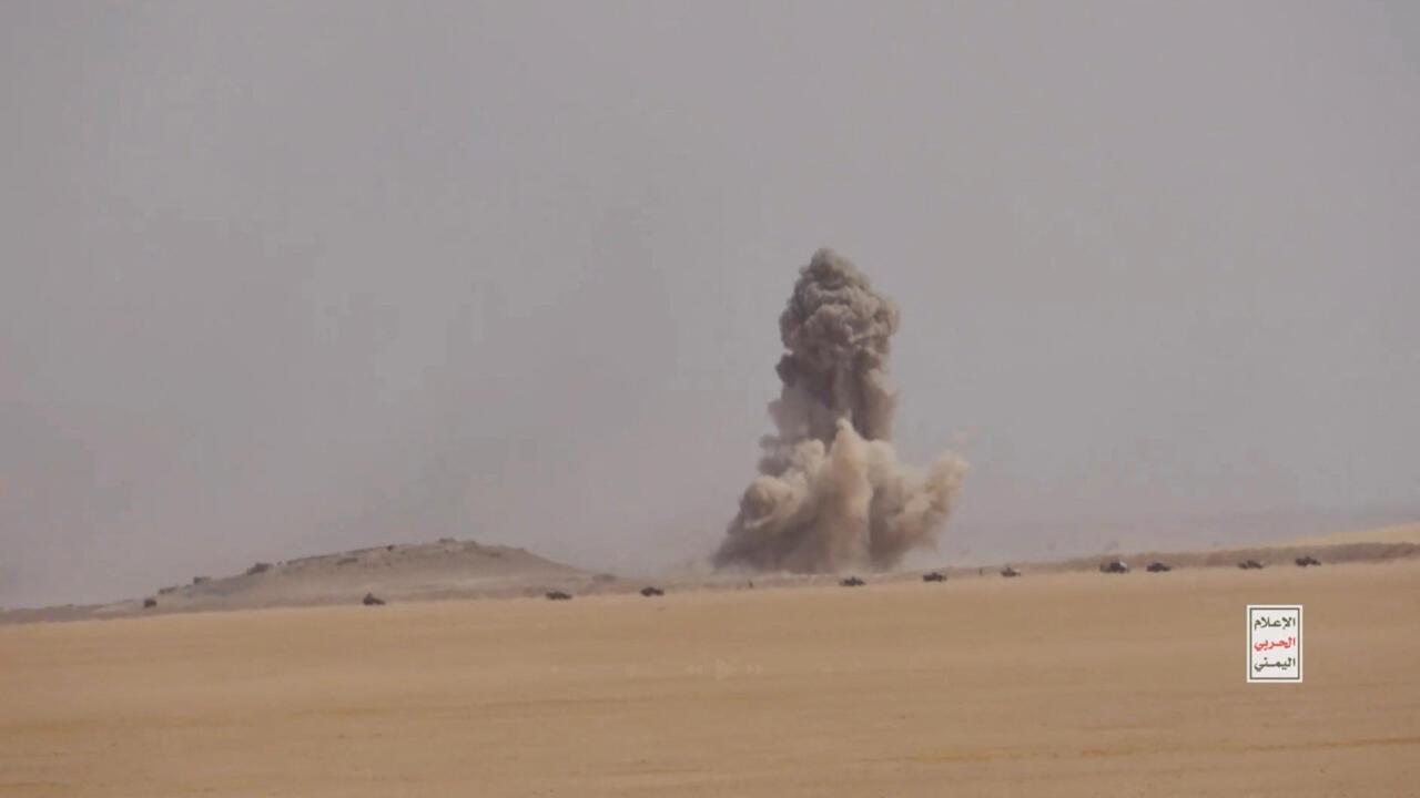 Dust plume rises from an apparent explosion in a barren desert landscape.