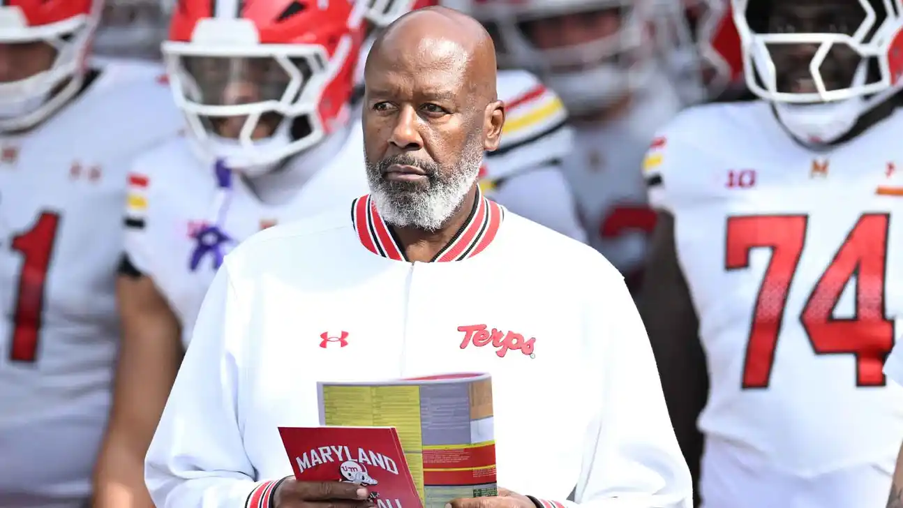 A man in a Maryland Terrapins jacket holds a folder in front of football players.