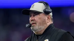 Bearded football coach wearing a cap and headset stands on the sideline during a game