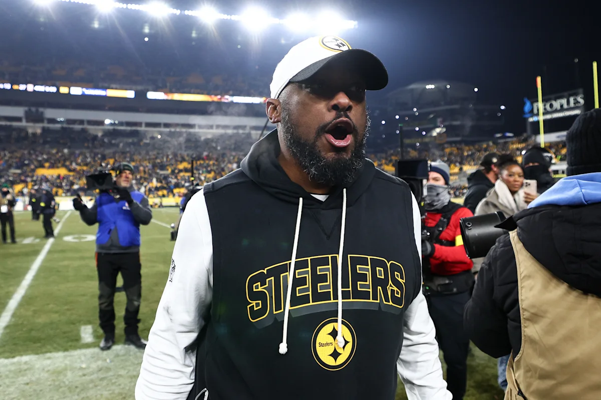 Man in Steelers hoodie and cap stands on a football field during a nighttime game.