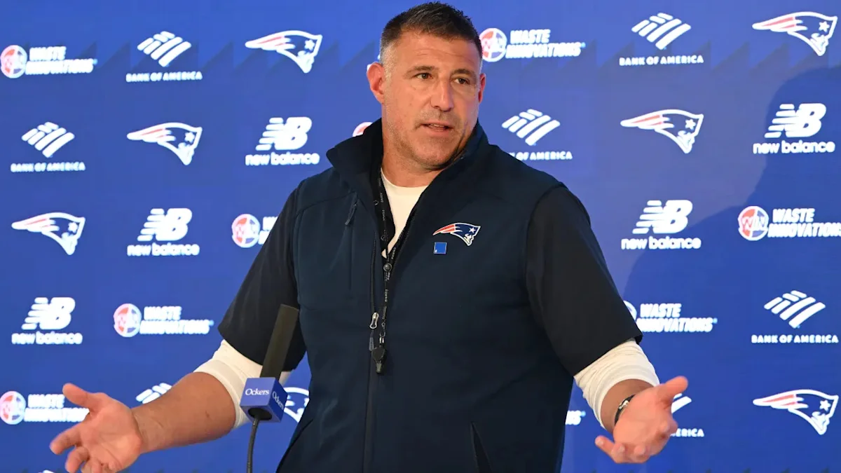 Man in Patriots vest gestures with both hands during a press conference.