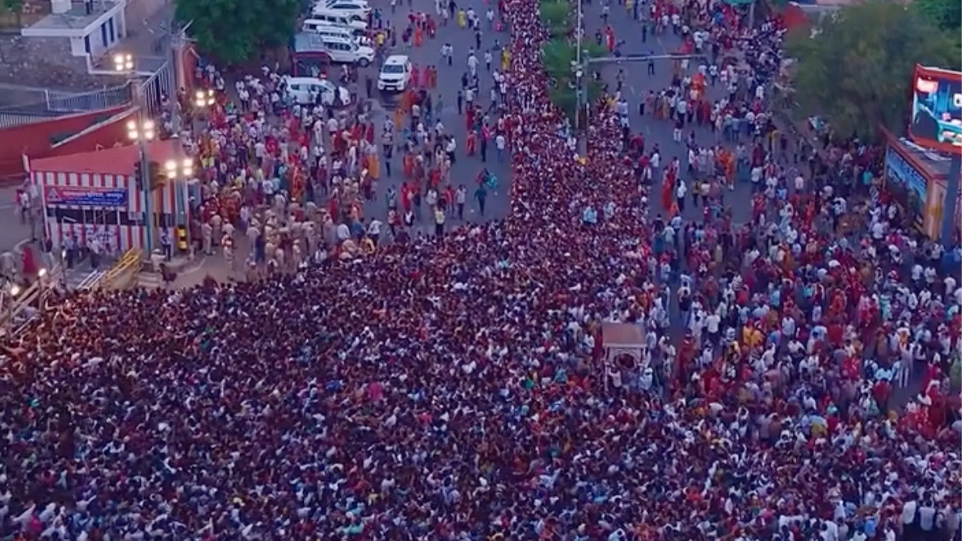 A huge crowd fills a broad street near temple buildings, with police and vehicles visible.
