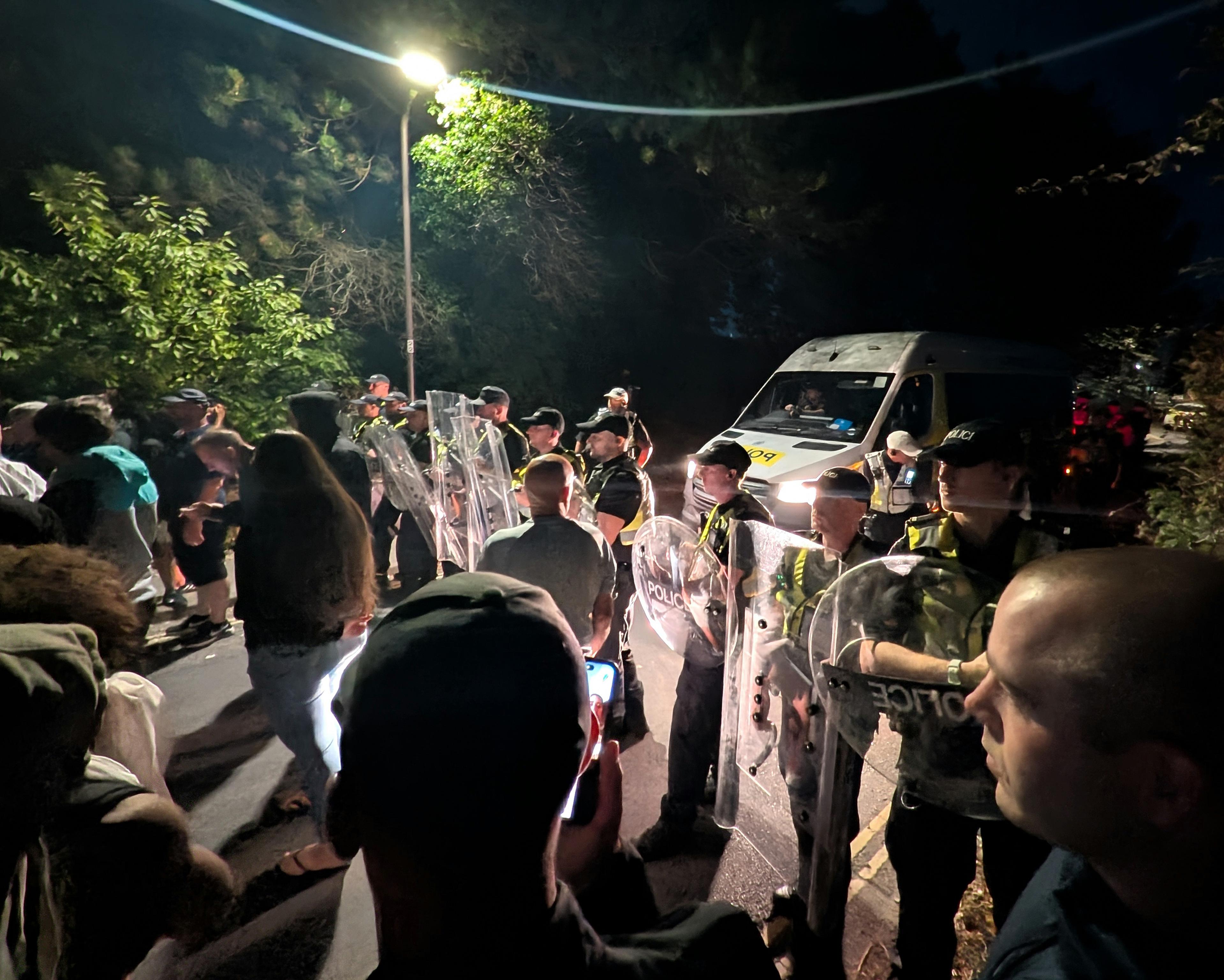 Police officers with riot shields face a crowd beside a police van at night.