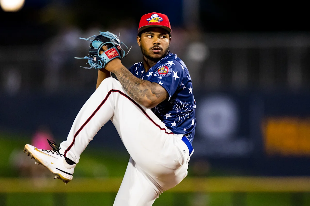 Baseball pitcher in a blue star-patterned jersey raises his leg during a nighttime pitch.