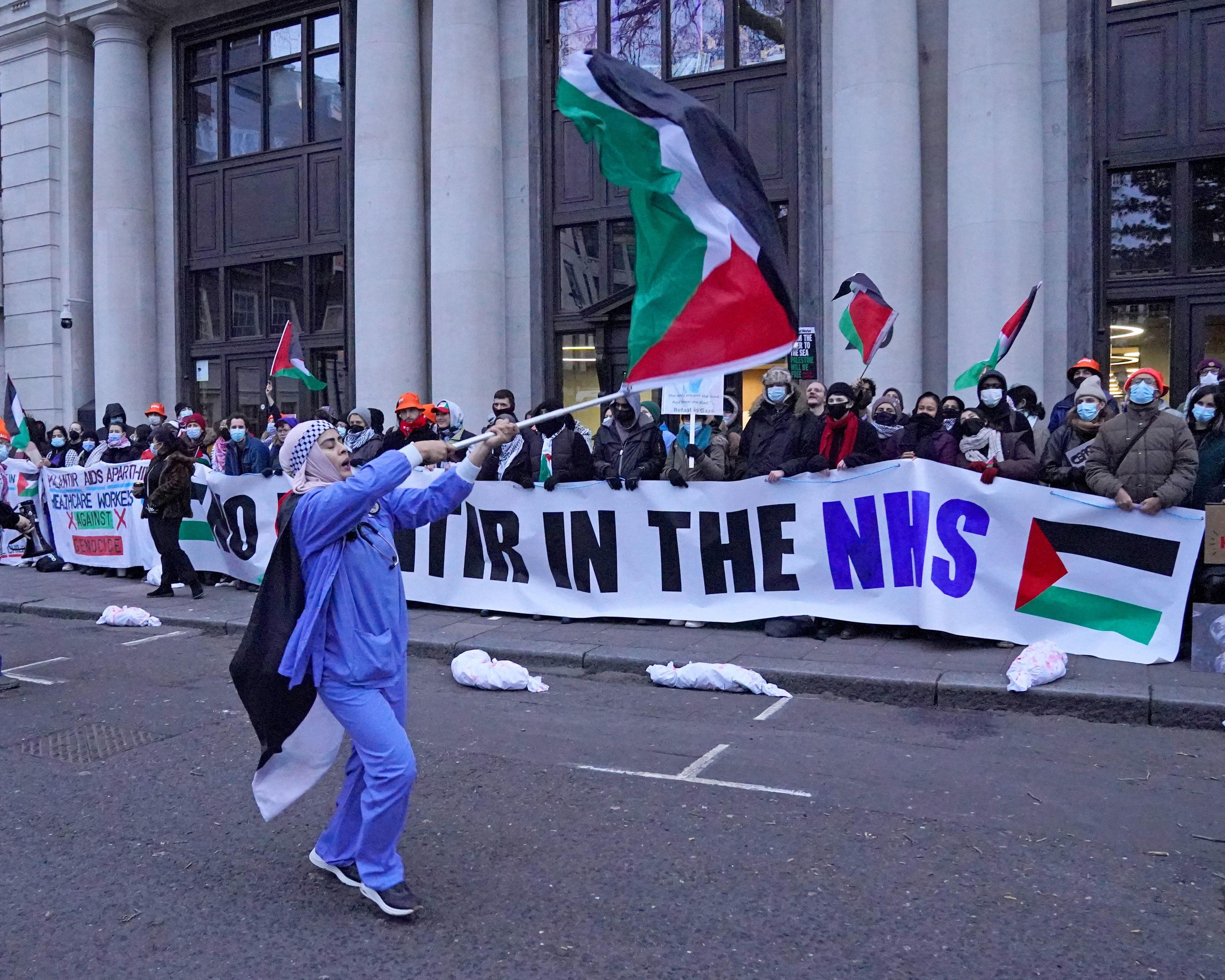 Protesters hold Palestinian flags and an NHS banner outside a columned building.