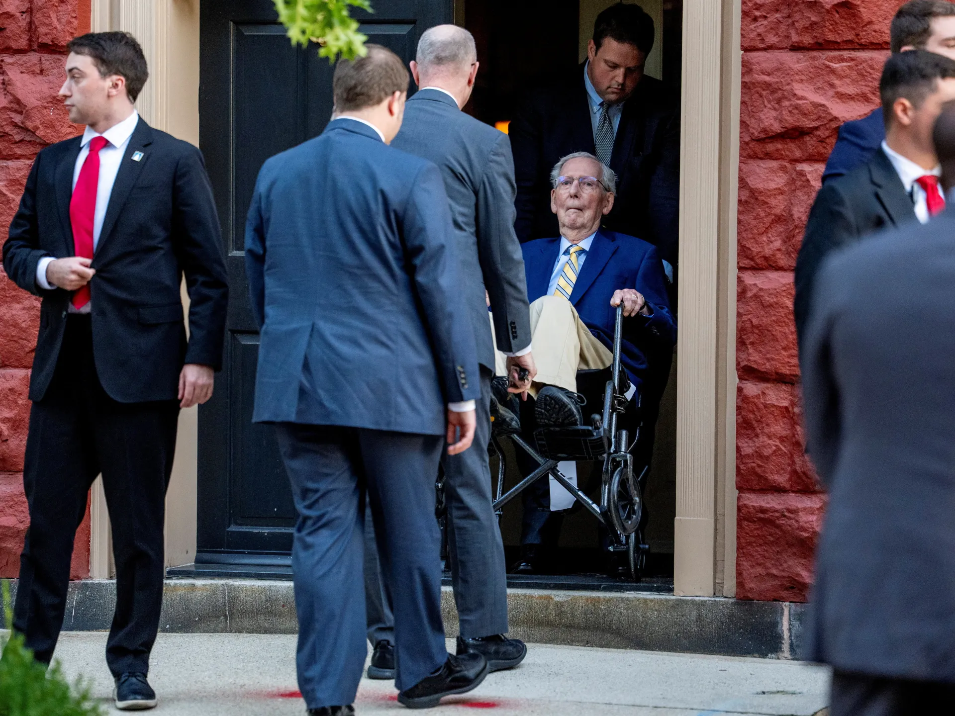 Mitch McConnell sits in a wheelchair near a doorway as aides and security personnel surround him.