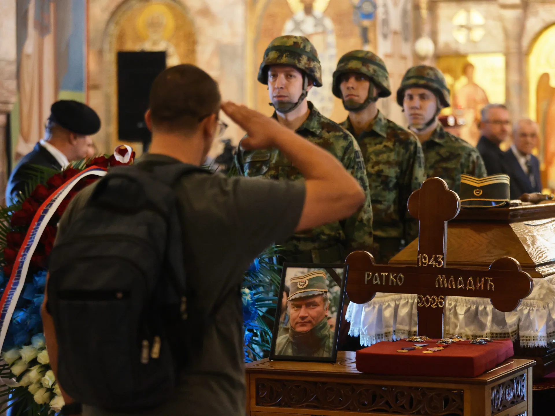 Mourners salute uniformed soldiers beside a coffin and memorial cross during a funeral ceremony.