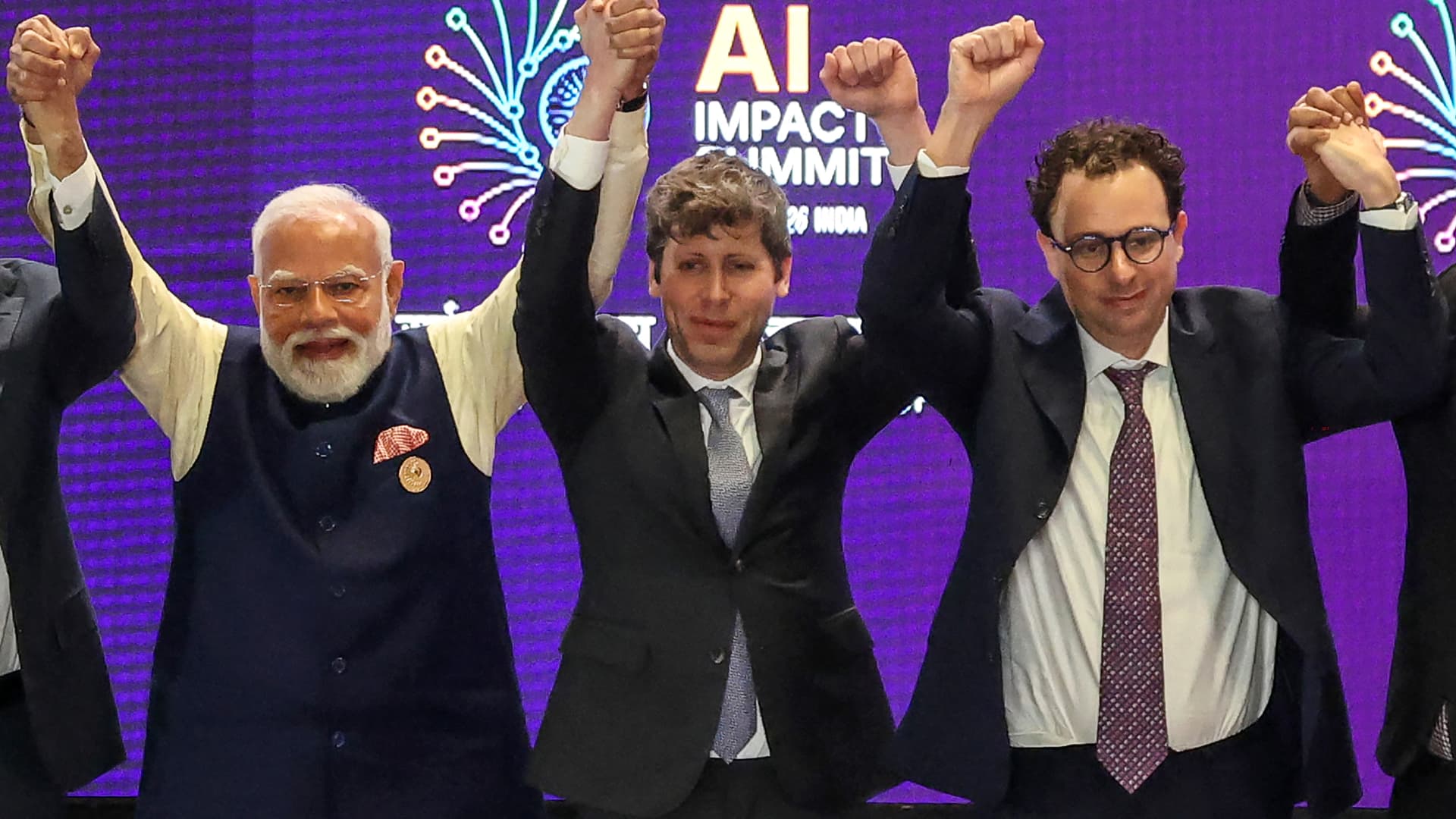 Three suited men raise clenched fists onstage before a purple AI Impact Summit backdrop.